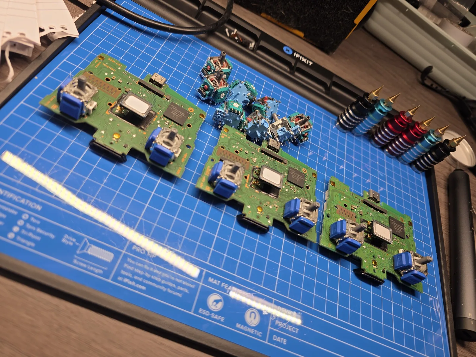 Three PlayStation controller boards with new TMR stick modules installed, laid out on a repair mat beside a pile of removed sticks and soldering tips on stands.